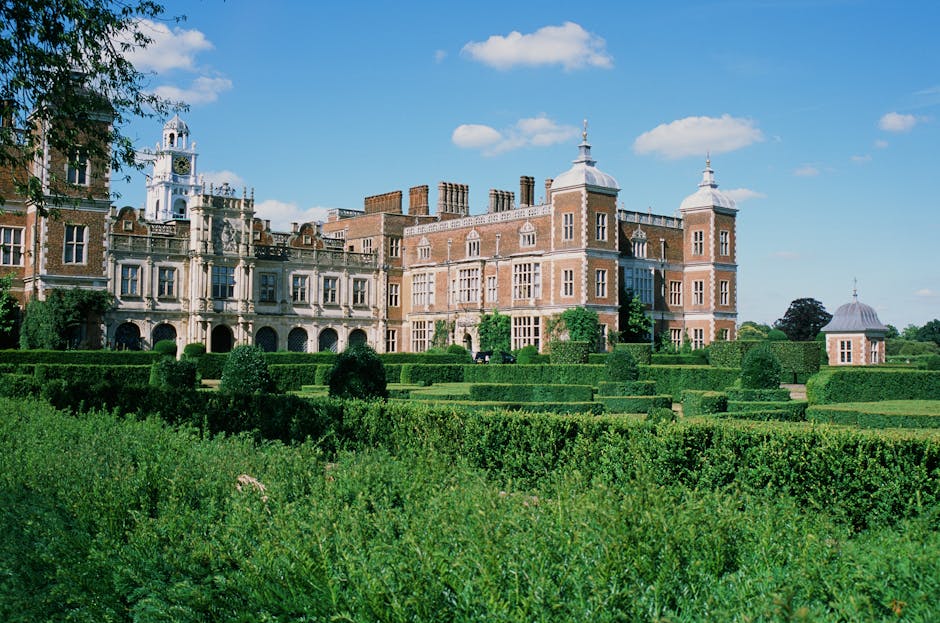 A historic castle with ornate architectural details and multiple turrets, surrounded by meticulously maintained formal gardens featuring neatly trimmed hedges, geometric flower beds, and lush green la