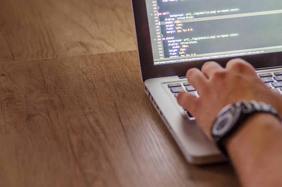 A person's hand, wearing a wristwatch, is typing on a laptop keyboard placed on a wooden surface. The screen displays lines of code with syntax highlighting, indicating programming or web development